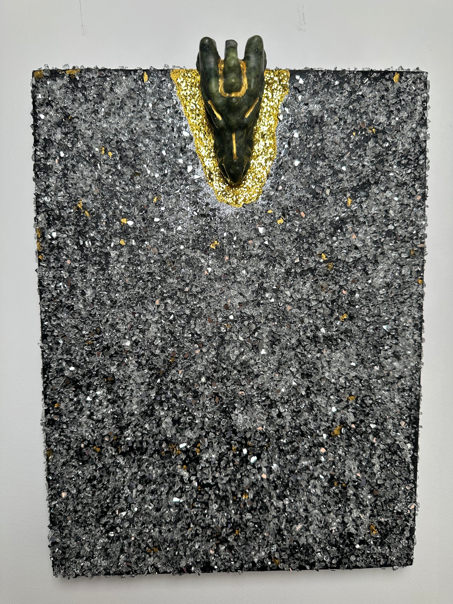 A square textured black and gray stone-like plaque or tile with scattered metallic flecks, featuring a small dark figurine placed on a gold glittery curved section along the left side, photographed against a plain light background.