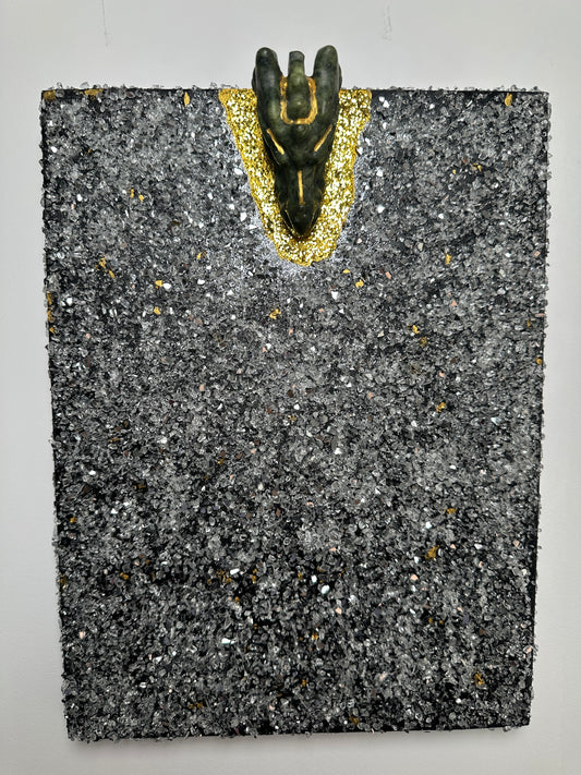 A square textured black and gray stone-like plaque or tile with scattered metallic flecks, featuring a small dark figurine placed on a gold glittery curved section along the left side, photographed against a plain light background.