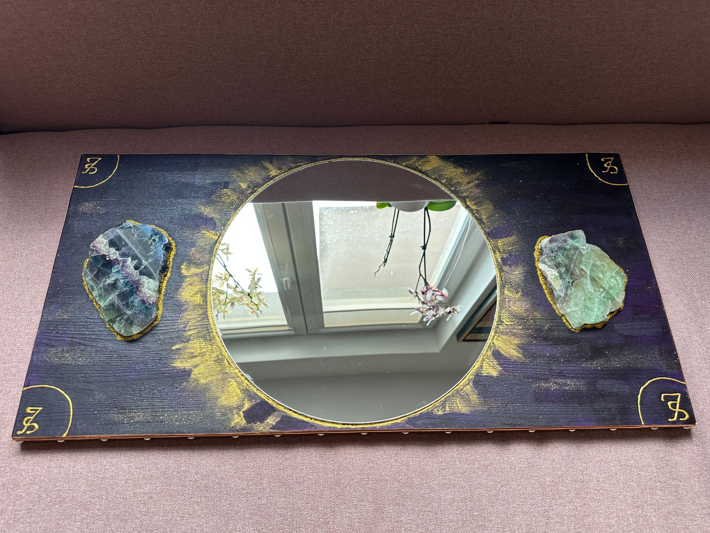 A rectangular decorative mirror with a dark navy and gold distressed background, featuring two raw green geode crystals on either side of the circular mirror center and gold corner symbols.