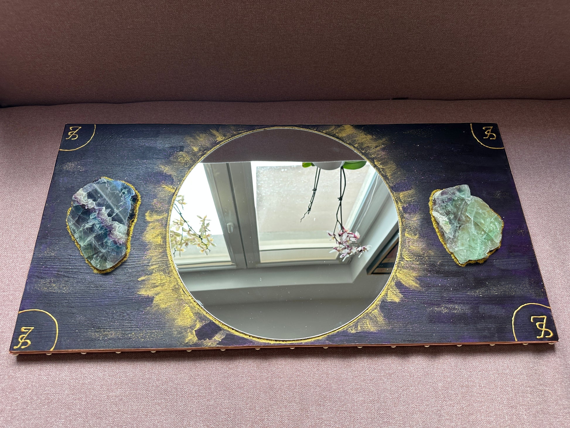 A rectangular decorative mirror with a dark navy and gold distressed background, featuring two raw green geode crystals on either side of the circular mirror center and gold corner symbols.